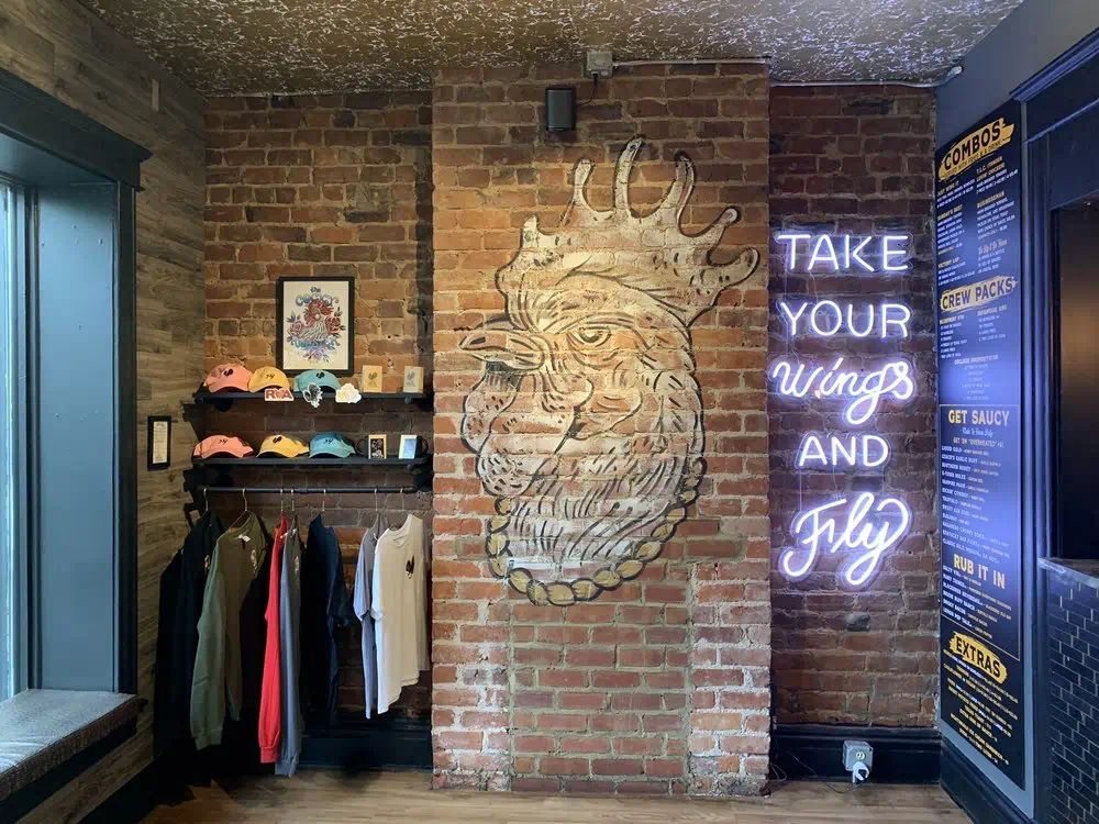 Merch wall with caps and shirts beside the brick rooster mural and neon sign